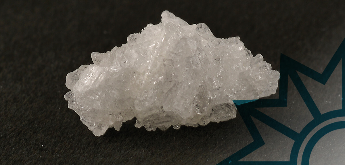 Nahaufnahme einer kristallinen Form von Crystal Meth auf dunklem Hintergrund. Die weißen, eisähnlichen Kristalle sind deutlich erkennbar und reflektieren das Licht.