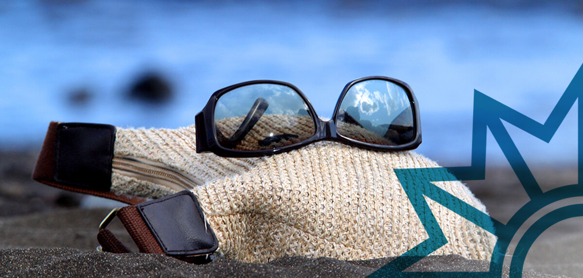 Nahaufnahme eines Strandes mit Sonnenbrille und Handtasche im Vordergrund. Im Hintergrund ist verschwommen das blaue Meer zu sehen.